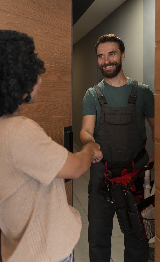 A smiling male technician wearing dark overalls and carrying a tool bag stands at the entrance of a home, shaking hands with a woman who is welcoming him. The setting suggests a professional service interaction.