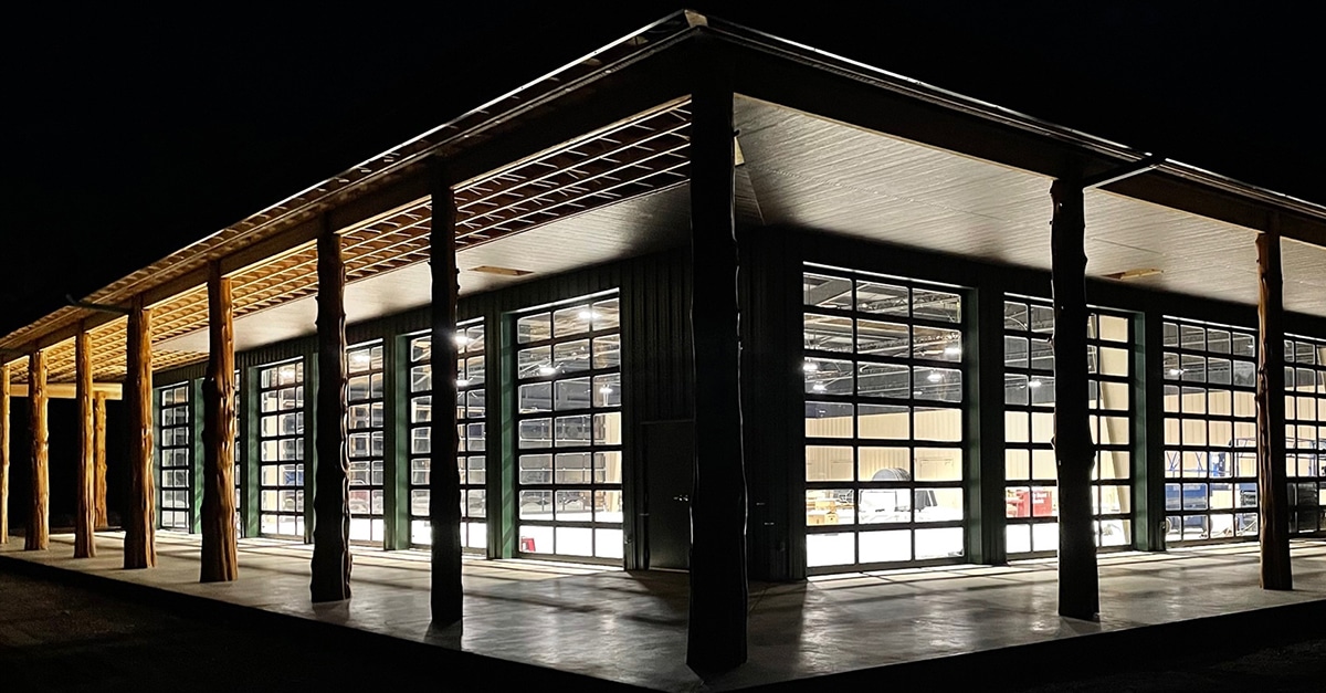 The image showcases a commercial building at night, illuminated from within, featuring C.H.I. Commercial full-view aluminum overhead doors. These doors have a grid of large glass panels, allowing clear visibility of the brightly lit interior. The building’s exterior design includes exposed natural wooden support columns and a wide overhang, adding a rustic yet modern aesthetic. The structure appears to be functional for commercial or industrial purposes, such as a workshop or showroom.