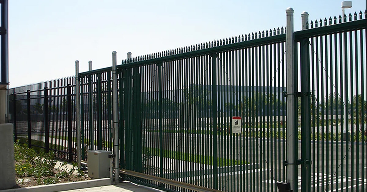 This image shows an industrial-grade black sliding gate with vertical bars, designed for enhanced security and durability. The gate features pointed tops to deter climbing, and it is supported by metal posts and an automated mechanism for smooth sliding operation. The setting appears to be a commercial or industrial facility, with a clear view of a parking lot and landscaping in the background. A warning sign is visible, indicating restricted access or safety precautions.