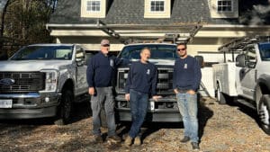 Picture of three Affordable Garage Door technicians dressed in their dark blue company sweatshirts standing in front of gleaming white company trucks.