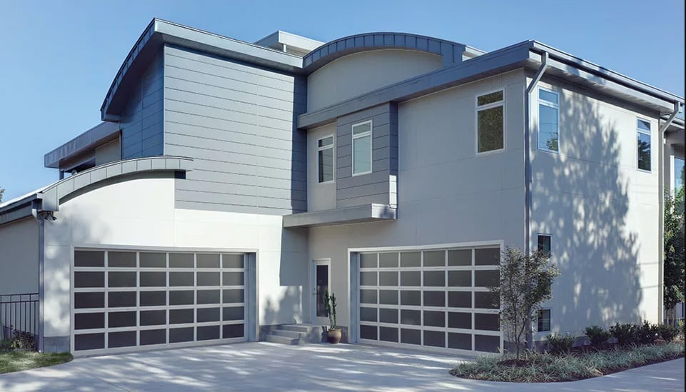 The garage doors by Amarr in this image showcase a contemporary design with a clean and minimalist aesthetic. They feature large, frosted glass panels framed in sleek, metallic aluminum, creating a modern grid pattern. The glass offers privacy while allowing diffused natural light to enter the garage. The symmetrical layout of the panels complements the home's overall architectural design, characterized by its sharp angles and neutral color palette. These Amarr doors enhance the home's modern appeal, blending functionality with cutting-edge style.