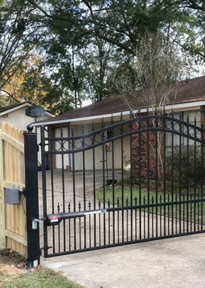 The image features the Amazing Gates Concord Swing Driveway Gate, designed for residential use. The gate is made of black metal with a decorative arched top and circular accents, combined with pointed finials at the bottom for added security. It is installed on a concrete driveway with a wooden fence on one side. The gate while functional and moderately stylish, is in a setting where the surrounding house and landscape could benefit from aesthetic enhancements to complement the gate's design.