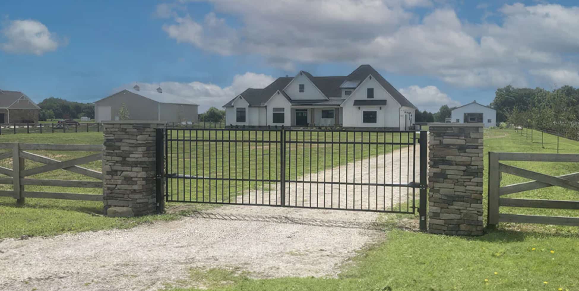 The image depicts a residential driveway entrance featuring the Telluride Swing Driveway Gates by Amazing Gates. The gates are made of black metal, framed by stone pillars on either side, with wooden fencing extending outward. A gravel driveway leads to a modern white farmhouse-style home, with a barn and additional outbuildings visible in the background, surrounded by open green fields under a partly cloudy sky. The scene emphasizes a balance of security, elegance, and rural charm.