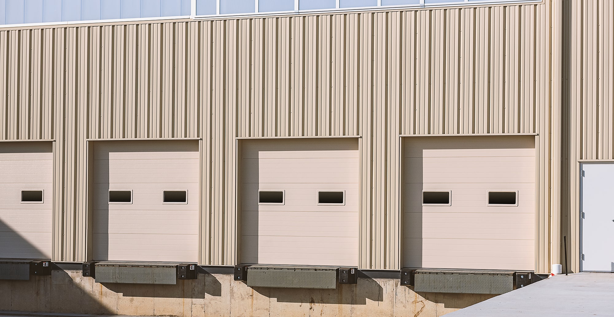 The image shows the exterior of a beige industrial building featuring three loading dock doors with small rectangular windows and protective dock bumpers. The structure is made of vertically ribbed metal panels, and there is a concrete foundation below the doors. A white personnel door is located on the far right. The setup is designed for truck loading and unloading.