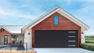 The garage door in this image is a sleek, modern design featuring a solid black finish with four horizontally aligned rectangular windows positioned on the right side. These windows provide a subtle geometric accent and allow for natural light while maintaining privacy. The minimalist style of the garage door complements the warm wood paneling of the surrounding gable and the overall clean lines of the house. This contemporary choice harmonizes well with the exterior's combination of natural and industrial elements.