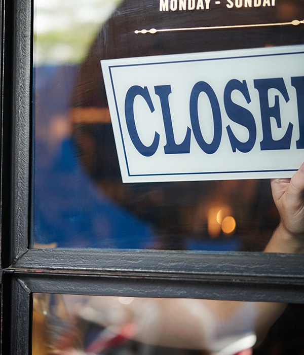 The image shows a person holding a "CLOSED" sign against the inside of a glass door or window. The reflection of the surroundings is visible on the glass, and part of the operational hours ("Monday - Sunday") is displayed above. The scene suggests a business closing for the day or temporarily not in service.