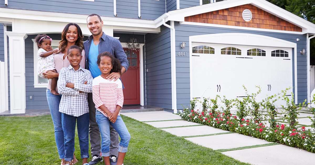 The image shows a smiling family of four standing outside a well-maintained suburban home with a prominent garage door in the background. The bright and welcoming setting emphasizes the importance of home and garage door safety, aligning with the article's title, "10 Must-Know Garage Door Safety Tips for Stafford Homeowners."