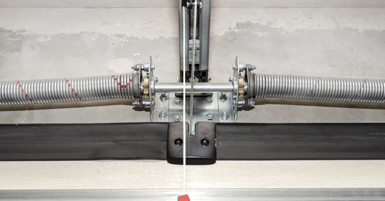 Close-up view of a garage door torsion spring system, showing two coiled metal springs mounted on a central shaft with brackets and fasteners. A metal cable runs vertically from the mechanism, attached to a red pull handle. The setup is installed on a concrete wall above the garage door.