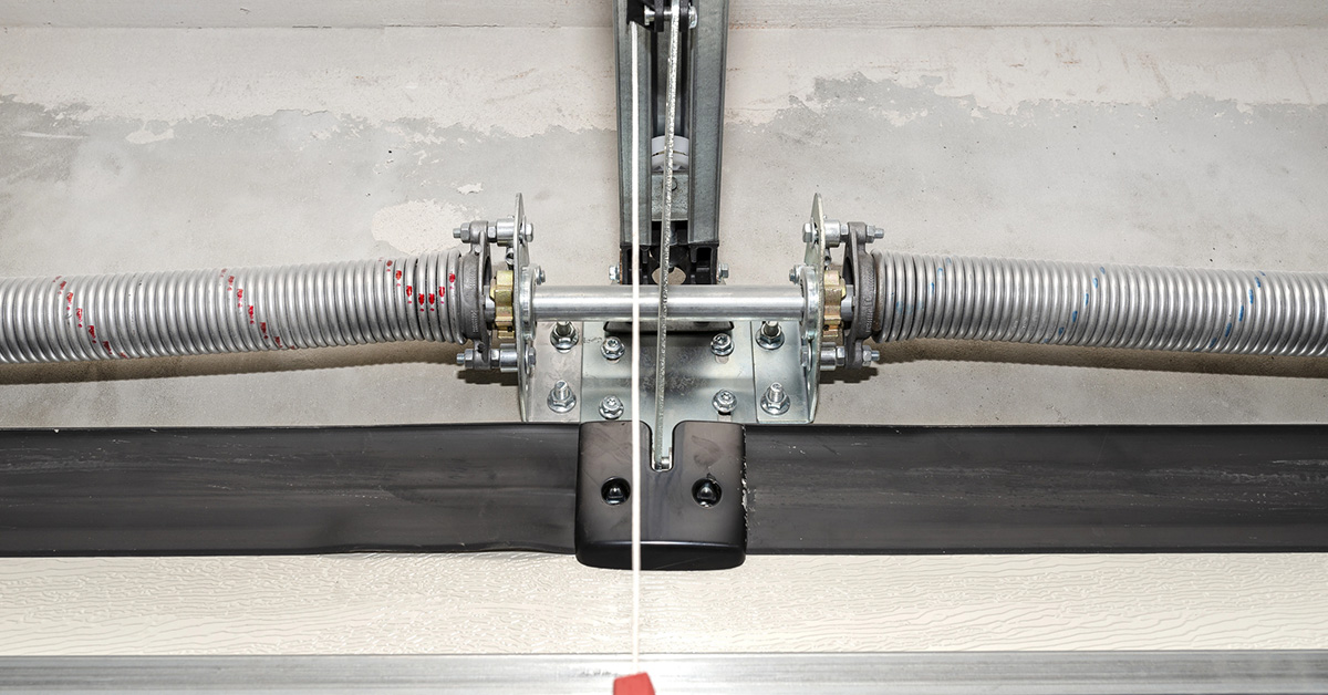 Close-up view of a garage door torsion spring system, showing two coiled metal springs mounted on a central shaft with brackets and fasteners. A metal cable runs vertically from the mechanism, attached to a red pull handle. The setup is installed on a concrete wall above the garage door.