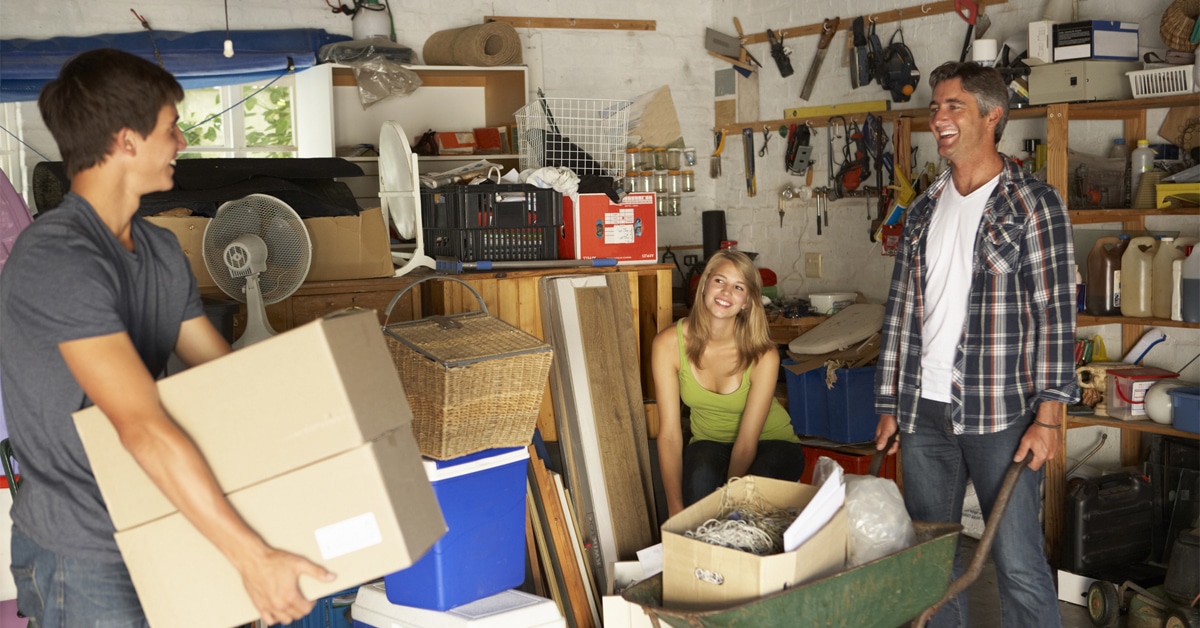 A group of three people—two men and one woman—are organizing a cluttered garage. The younger man in the foreground is carrying two cardboard boxes, while the woman is sitting on a stool, smiling. The older man, wearing a plaid shirt, is holding a wheelbarrow filled with miscellaneous items. The garage contains various tools, storage bins, and equipment. This setting suggests a home improvement project, possibly including a task like garage door spring repair among their organizing efforts.