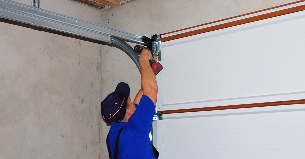 A technician in a blue uniform and cap is installing or repairing a garage door mechanism using a power drill. The garage door is white with wooden trim, and the metal tracks are mounted on the ceiling and wall. The background consists of an unfinished concrete wall.