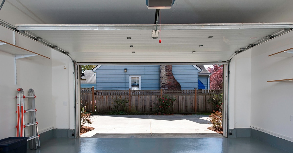 View from inside a garage with an open door, showing a driveway leading to a fenced backyard with a blue house in the background. The image highlights the need for garage door spring repair in Stafford, as properly functioning springs are essential for smooth operation and safety.