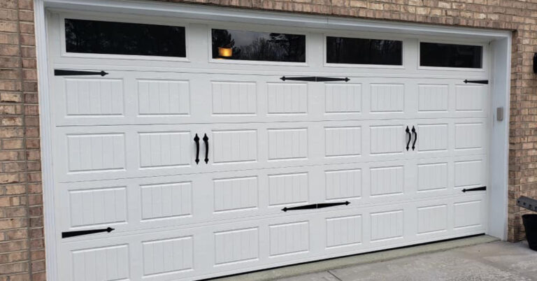 White sectional garage door with decorative black hardware and four small windows at the top, set in a brick exterior.