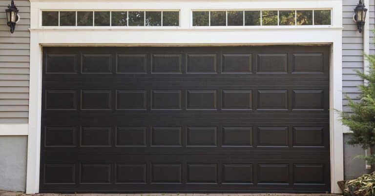 Closed black garage door on a residential home, framed by white trim and small upper windows, illustrating the type of door that may require repair.