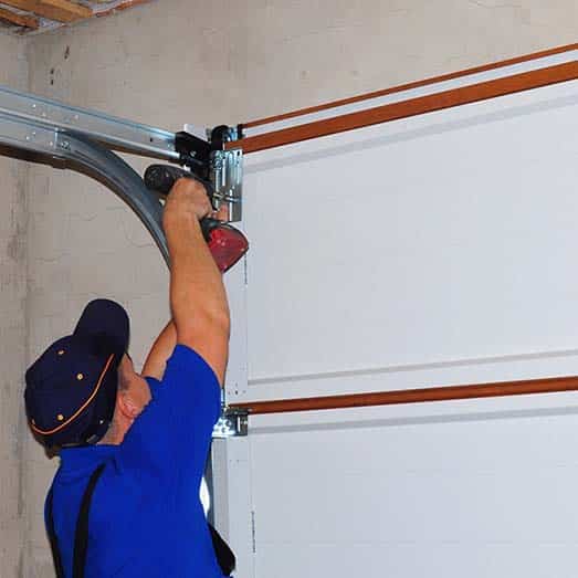 A technician in a blue uniform and cap repairing a garage door mechanism using a power drill. The garage door is white with wooden trim, and the metal tracks are mounted on the ceiling and wall. The background consists of an unfinished concrete wall.