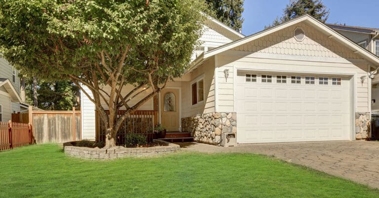 uburban cream house with two-car garage, stone trim, tree-shaded lawn, and front porch.