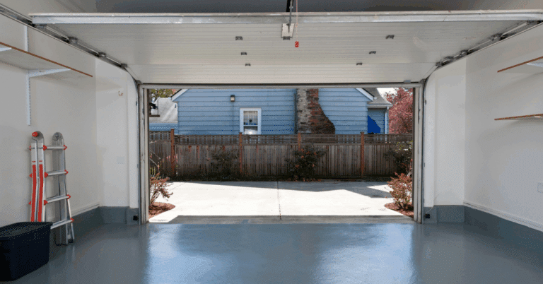 Clean, empty garage with ladder and shelves, door open to driveway and blue house.